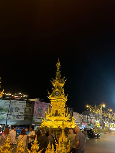 The image displays the illuminated Golden Clock Tower (Ho Nalika Chalermprakiat) at night, situated at the intersection of Phaholyothin Road and Jet Yod Road in Chiang Rai City Municipality, Chiang Rai, Thailand. The central subject is an ornate, multi-tiered clock tower constructed in a golden Thai architectural style, brightly lit. Approximately 10-15 individuals, adults in casual clothing, are observed gathered around the base of the tower, appearing to observe it. Ornate golden decorative elements are in the immediate foreground. In the background, multi-story urban buildings display illuminated signage, some with Thai script and partial English text, lining the street. A street is visible to the right of the tower, occupied by at least one dark-colored vehicle. Decorative string lights are strung across the street on the right, contributing to the urban nighttime illumination. The sky is dark.