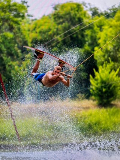 A man is wakeboarding. He is shirtless, wearing blue swim trunks. He is in the air, holding onto a tow rope attached to a wakeboard. Water is spraying around him. Green trees are in the background. The image was taken in Bacong, Philippines.