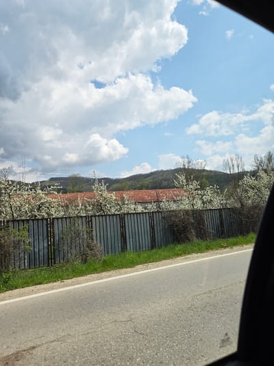 The image shows a view from a vehicle, looking across a road at a fence, a building, and a hillside. The sky is visible in the upper portion of the image, with both blue sky and cumulus clouds. The road is gray with a white line marking the edge and a strip of green grass separating it from the fence. The fence is metallic and appears to be made of corrugated sheets. Behind the fence, there is a building with a red tile roof, beyond which a tree-covered hillside rises. The white blossoms on the trees suggest springtime. The image was taken in Dobrodan, Bulgaria.