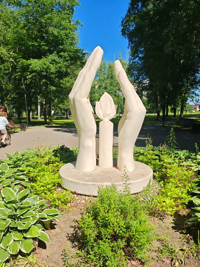 This image depicts a public park in the city of Buy, Russia, featuring a white sculpture of two cupped hands surrounding a candle flame atop a cylindrical base. The sculpture is situated in a garden bed filled with hosta plants with variegated green and white leaves and various other green shrubs and plants. In the background, a paved walkway is visible, with a park bench and a trash receptacle to the left. A person on a bicycle, wearing a light-colored shirt and shorts, is riding on the walkway. Beyond the walkway, a line of trees with lush green foliage stretches across the scene, and a clear blue sky is visible above.