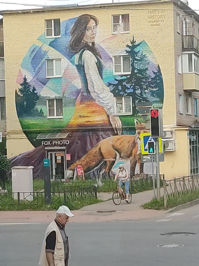 This image captures a street scene in Staraya Russa, Russia, featuring a multi-story building adorned with a large mural. The building, constructed of light yellow brick, has multiple windows integrated into the mural.

The mural covers the majority of the building's facade. Its upper portion depicts a woman with long dark hair, facing towards the viewer's left. She wears a light-colored long-sleeved garment with a darker, sleeveless garment over it. Behind her, abstract geometric shapes in shades of blue and purple transition into a landscape featuring evergreen trees and a sunset/sunrise-like gradient of greens, oranges, and yellows. In the top-right corner of the mural, faded text reads "PAGES OF HISTORY," "ALLAGEYEV," and "MANOS."

At the base of the mural, a large, recumbent fox-like animal is depicted, its brown and tan fur prominently rendered. Directly beneath the fox and centrally positioned at street level is a commercial establishment named "FOX PHOTO," indicated by black lettering above its entrance.

On the street and sidewalk below:
*   In the immediate foreground, a man walks from right to left across the asphalt road. He wears a white baseball cap, a light beige utility vest over a dark short-sleeved shirt, and dark pants.
*   On the concrete sidewalk to the right, a woman cycles away from the viewer on a bicycle with a red frame. She wears a light-colored t-shirt, blue shorts or capris, and a light-colored visor.
*   A pedestrian traffic light pole is positioned at the edge of the sidewalk and road. The upper traffic signal displays a red light, while the lower pedestrian signal shows a green walking figure. A blue square sign with a white triangle indicating a pedestrian crossing is attached to this pole.
*   A low, dark green metal fence runs parallel to the sidewalk, separating it from a strip of grass and shrubs.
*   A gray utility box or pillar stands near the "FOX PHOTO" entrance, beside a green pole that appears to have an "SOS" logo.
*   The asphalt road shows visible manhole covers.
*   The adjacent section of the building to the right features a ribbed facade, several windows, and enclosed balconies. Multiple outdoor air conditioning units are affixed to this section of the building.