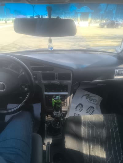 The image captures the interior of a vehicle from the driver's perspective, looking forward through the windshield into a brightly lit street scene.
**Vehicle Interior:** The dashboard is dark gray with silver accents around air vents and control panels. A black steering wheel with a visible emblem is on the left. The manual gear shifter is prominently featured in the center console, topped with a green "Gorilla Energy Drink" can acting as a custom knob. A white plastic bag with "Neo Professional" branding and two shoe print icons rests on the passenger side floor area. A rearview mirror hangs from the top center of the windshield, with a small, clear air freshener containing blue liquid suspended beneath it. The driver's left leg, clad in blue jeans, is visible on the lower left. The passenger seat, on the right, is dark fabric with a light-colored stitching pattern.
**Exterior Environment:** The street outside is illuminated by strong sunlight, creating glare on the windshield. The ground appears to be a light-colored paved surface. In the mid-distance, a large, ornate architectural structure, possibly a gateway or building entrance, with a distinct blue hue and yellow accents, is visible. To the right of this structure, a dark-colored sedan is parked or stopped with its driver's side door open. The sky is clear blue. The scene is located in the city of Pakhtakor, Uzbekistan.
