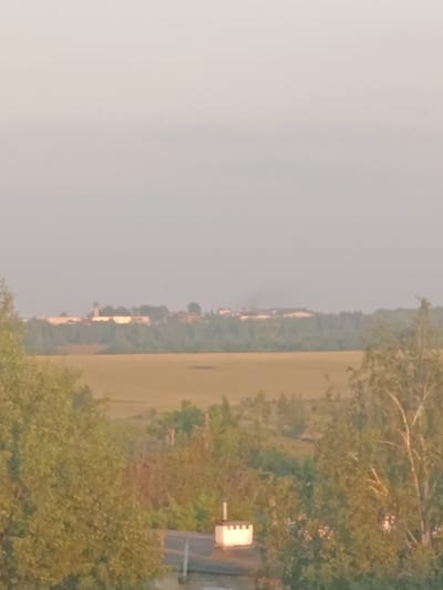 This image depicts a rural landscape, likely in Pobedinka, Russia, under a hazy, overcast sky. The time of day appears to be late afternoon or early evening, given the soft, diffused light.
In the foreground, the tops of several trees with green and yellow foliage are visible, suggesting a temperate climate during late summer or early autumn. On a rooftop, a small white structure with a decorative, crenellated top is present, hinting at residential buildings.
The middle ground is dominated by a wide, open field of dry, golden-brown grass or crops, stretching towards a line of trees. Beyond this, in the distance, a sprawling complex of buildings with a tall, tower-like structure is visible. This complex, possibly an industrial facility or a settlement, is partially obscured by the trees and haze. A faint plume of smoke rises from the complex, indicating some form of activity.
The overall impression is one of a quiet, expansive rural setting, with a hint of industrial presence on the horizon. There is no visible text in the image. The atmosphere is calm and somewhat subdued, consistent with the hazy weather and soft lighting.