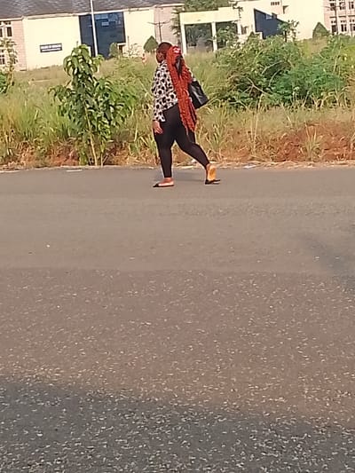 A woman is walking on a paved road in Ogboko, Nigeria. She is wearing a white and black patterned blouse, black leggings, and flat shoes. She has long, reddish-brown braids and is carrying a black purse on her shoulder. The road is gray and takes up most of the frame. To the side of the road, there is vegetation growing along the edge. Behind the woman and the vegetation, there are buildings in the background. One of the buildings has "College of Medicine" written on it.
