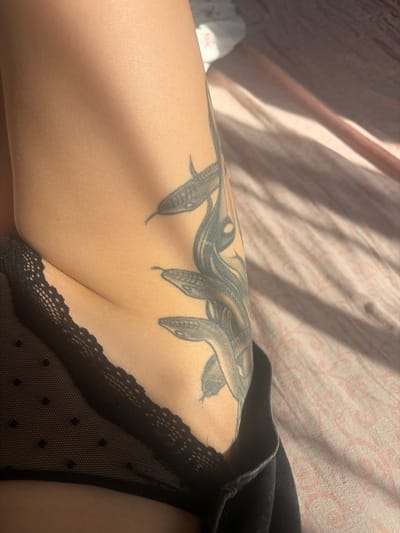 The image shows a close-up view of a person's leg and thigh, adorned with a tattoo of a snake-like creature with multiple heads. The person is wearing black lace underwear and dark clothing. The background consists of a pink patterned sheet, partially illuminated by sunlight. The image does not contain any information about the city of Shlisselburg, Russia.