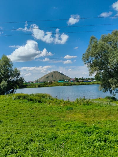 This is a wide-angle, eye-level photograph of a pastoral landscape in the city of Yuraktau, Russia. The scene is dominated by a tranquil body of water in the middle ground, reflecting the bright blue sky dotted with fluffy white clouds. To the left and right of the water are lush green grassy banks, with one bank on the left featuring a large, leafy tree. Across the water, a row of small houses with red and blue roofs forms a village nestled against the base of a conical, verdant hill that rises in the distance. The overall impression is one of a serene rural setting on a clear, sunny day.