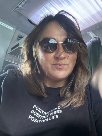A woman is taking a selfie on what appears to be public transport. She is wearing sunglasses and a black t-shirt with white text that says "POSITIVE MIND POSITIVE VIBES POSITIVE LIFE." The background shows the interior of a vehicle, including seats and overhead compartments. There is also a sticker on the wall with Russian text that translates to "maximum load 30 kg."