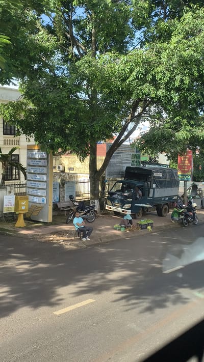 A sun-drenched street scene in Trung Hồ, Vietnam shows a light blue truck parked on the side of the road, its cargo area covered with a grey tarp. A man is seated in the driver's seat. Two women are crouched beside the truck on the sidewalk, appearing to be selling produce. One woman is sorting green fruits, possibly watermelons or melons, from a basket, while the other is attending to a pile of durians.
Adjacent to the truck, a motorcycle is parked. Further down the sidewalk, a man in a light blue shirt and dark pants sits on a white plastic chair. A yellow mailbox with "BƯU ĐIỆN" (Post Office) and "THU TIỀN ĐIỆN" (Electricity Payment) written on it is positioned near the man. Behind the mailbox, a signpost with multiple rectangular panels listing services is visible, including "CHỢ VAY NHANH TIÊU DÙNG" (Fast Consumer Loans) and "ĐIỂM BÁN HÀNG TIỆU DÙNG" (Convenience Store).
The background features a cream-colored building with dark-trimmed windows, shaded by a large tree with abundant green foliage. Signs with Vietnamese text are attached to the building and a fence. The road in the foreground displays a yellow painted line. Shadows from the tree are cast across the road and sidewalk.