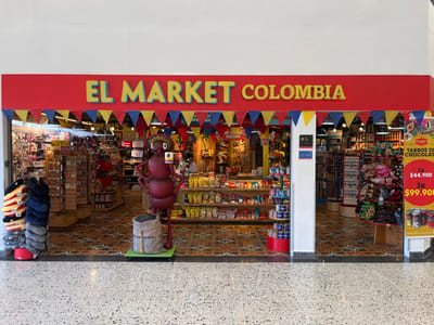 The image displays the brightly lit storefront of a retail establishment named "EL MARKET COLOMBIA," situated within an indoor commercial area in the city of Girón, Colombia. The store's facade is red, featuring the name in yellow lettering with blue borders, and is adorned with a decorative string of red, yellow, and blue triangular pennants.
Two open entrances, separated by a central white pillar, provide access to the store's interior, which is densely stocked with merchandise on shelves and racks. Prominently positioned in front of the left entrance is a large, reddish-brown anthropomorphic ant statue. This statue wears a straw hat and holds a small white object in its right hand while its left hand rests on its hip. At the base of the ant statue, a light-colored sack filled with dark, granular material is visible, bearing text that includes "BUCARAMANGA" and what appears to be "HORMIGA" or "HORMIGAS." To the far left, a stack of colorful folded textiles is displayed, alongside a dark, tall display stand with various small items.
Inside the store, a single individual is visible behind a counter on the right side. A large vertical sign on the right of the storefront advertises "TARROS DE CHOCOLATE" with prices listed as "1x $44.900" and "3x $99.900." The store's interior floor is covered with an intricate, colorful mosaic-like tile pattern, contrasting with the light-colored terrazzo flooring of the surrounding commercial space.