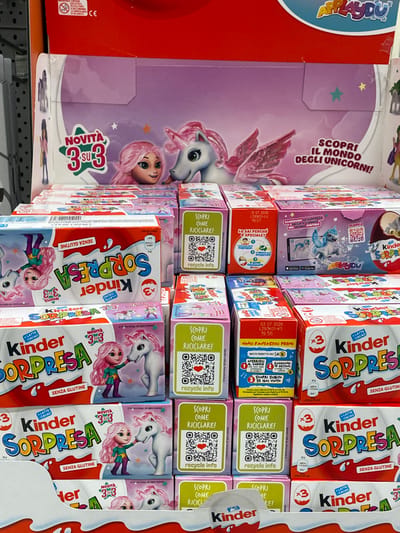 The image displays a retail store shelf in Monsummano Terme, Italy, featuring a tiered arrangement of Kinder Surprise and Kinder Applaydu products. The primary theme is unicorns and a female character with pink hair. A large cardboard header sign at the top showcases a pink-haired character looking at a white winged unicorn, with text in Italian reading "NOVITÀ 3 SU 3" (New 3 out of 3) and "SCOPRI IL MONDO DEGLI UNICORNI!" (Discover the world of unicorns!). Below this, multiple rows of Kinder Surprise boxes are stacked. White and red boxes labeled "Kinder Sorpresa" and "Senza Glutine" (gluten-free) are visible, some indicating "x3" for three eggs. Smaller individual boxes in pink, purple, and green are interspersed, many featuring "SCOPRI COME RICICLARE!" (Discover how to recycle!) with QR codes and "recycle info". Other boxes display text like "Scansiona la sorpresa nel mondo digitale" (Scan the surprise in the digital world) with an image of a phone showing a digital unicorn, and "Vinci fantastici premi!" (Win fantastic prizes!). The products are uniformly aligned, indicating a structured commercial display.