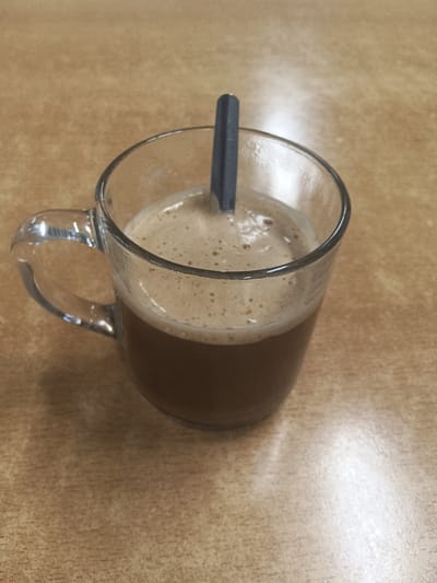 A close-up, overhead shot captures a clear glass mug filled with a frothy brown beverage, likely coffee or a similar drink. A dark grey stirrer rests within the mug. The mug is positioned on a light brown, wood-grain textured surface, possibly a table. The lighting casts a bright reflection on the table surface to the right of the mug. The background is out of focus. The image is taken indoors. No specific location in Sexbierum, Netherlands is depicted or implied.