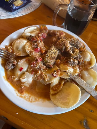 The image shows a plate of ravioli with a meat-based sauce, garnished with what appears to be grated cheese. A fork and knife are present. The dish is on a white plate placed on a wooden table. A glass pitcher with a dark liquid and a package of food are visible in the periphery. Based on the metadata, the image was taken in Villa General Belgrano, Argentina.