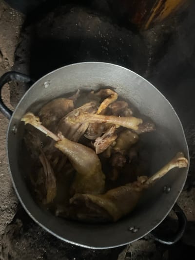The image shows a pot of cooked chicken pieces simmering in a dark, rustic setting. The pot, made of a dull metal, is filled with chicken legs and other parts, some of which appear to be intact while others are partially deboned. The chicken is a light yellow to brown color, suggesting it has been boiled or stewed. Wisps of steam rise from the pot, indicating that the food is hot and freshly cooked.

The pot is placed on a rough, earthy surface, possibly a traditional outdoor stove or fireplace, with scattered debris visible around it. The background is dark and indistinct, with hints of rough textures and perhaps foliage or wooden elements. The lighting is dim, suggesting it could be nighttime or an indoor space with low light. There is no visible text in the image.

The scene evokes a sense of traditional cooking methods in a rural environment, possibly in Aiyegunle, Nigeria, as suggested by the location context. The focus is solely on the food and the cooking vessel, with no people or other significant objects visible. The overall impression is one of a simple, hearty meal being prepared.