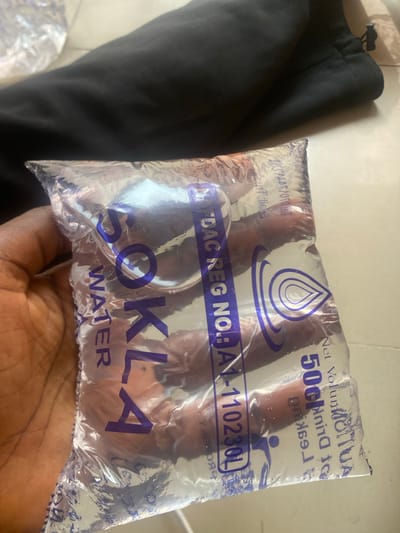 A person's hand is holding a clear plastic sachet of water. The sachet is filled with water and has purple text and graphics printed on it. A black piece of fabric, possibly clothing, is visible to the left of the sachet. In the background, the surface appears to be a light-colored tiled floor. The image has a bright and natural light source, suggesting it was taken during the daytime, possibly outdoors or in a well-lit indoor space. The overall scene is simple and focuses on the water sachet.

Visible text on the sachet includes:
- "SOKLA"
- "WATER"
- "Net Volume 50cl"
- "DO NOT DRINK TO LEAKAGE"
- "NAFDAC REG. NO: NA1-110230L"