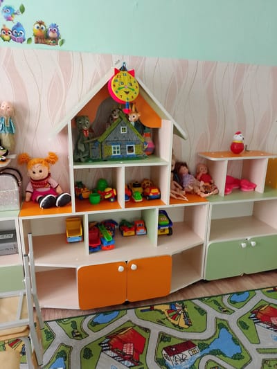 A toy house bookshelf with various toys is shown in an indoor setting. The bookshelf is white with some orange and green accents. On the top shelf, there is a decorative clock shaped like a cat's face, a miniature house with toy animals, and other small toys. The middle shelf features several dolls, and a few toy cars. The bottom shelf is mostly empty with a few more toy cars visible. The background consists of a wall with patterned wallpaper and a light blue upper section. A rug with a city or road design is visible on the floor. There are no people present in the image. The image is taken from a slightly elevated angle. The location of this image is not specified in the provided information, and it is not possible to determine if it is in Zhirnovsk, Russia.