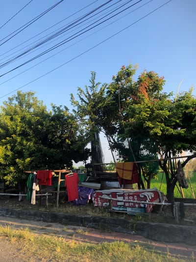 The image depicts an outdoor scene with several items of clothing hanging on a makeshift drying rack. The rack appears to be constructed from bamboo poles and is situated in front of lush green trees. Some of the clothing items are brightly colored, including red and yellow garments. In the background, a field of green crops is visible, possibly rice paddies, suggesting a rural or semi-rural setting. The sky is clear and blue, with power lines running diagonally across the upper portion of the frame. The foreground shows a paved area, possibly a road or sidewalk, with some dry grass and dirt. The overall impression is one of a humble dwelling or yard in the city of Kalidawir, Indonesia.