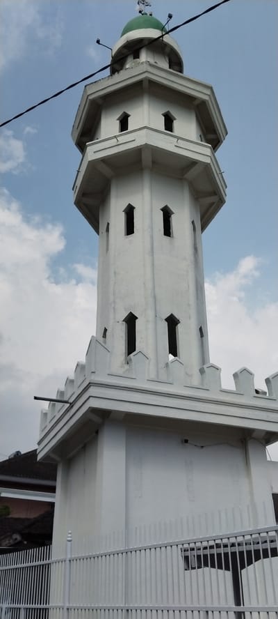 This is a ground-level, vertical, outdoor, eye-level photograph of a white minaret with a green dome, located in the city of Mataram, Indonesia. The minaret is octagonal in shape and features several tiers. The lower portion of the minaret is wider and has a crenelated parapet. Above this, the minaret narrows and has a series of pointed arch-shaped windows on each level. The top of the minaret is capped with a green dome, topped with a finial.
Several black electrical wires are visible in the upper portion of the image, running diagonally across the sky. Two black spotlights are mounted on the upper portion of the minaret, just below the dome. A white metal fence with vertical bars is in the foreground, partially obscuring the base of the minaret. Behind the minaret and to the left, a portion of a building with a dark-tiled roof is visible. The sky is a pale blue with scattered white clouds. No people are visible in the image. The overall impression is one of a religious structure under a partly cloudy sky.