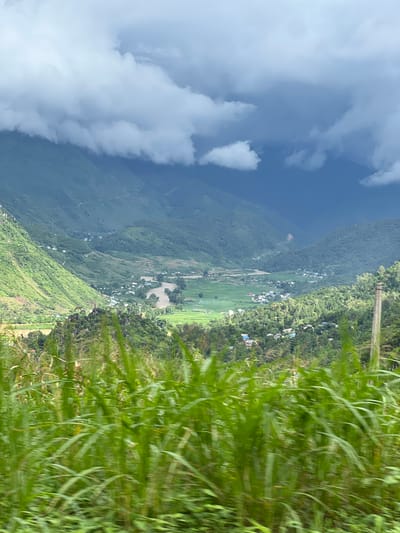 A wide, scenic vista captures a valley in Lùng Tám Commune, Vietnam, under a dramatic sky. The foreground is dominated by blurred green foliage, likely tall grass or reeds, suggesting movement, perhaps from a vehicle. Beyond this, the mid-ground reveals a lush green valley dotted with small houses and rice paddies, a winding river cutting through the landscape. The valley is framed by imposing, verdant mountains that ascend towards the background. The sky above is a striking contrast of bright, fluffy white clouds on the left, transitioning to dark, ominous storm clouds on the right, with a sliver of bright light breaking through in the distance. The scene depicts a rural, mountainous environment with signs of human habitation within a natural, atmospheric setting.