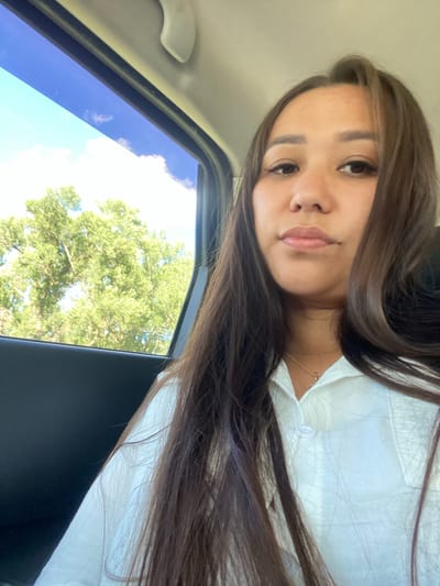 A young woman with long, dark brown hair is captured in a selfie from inside a vehicle. She is wearing a white collared shirt and a delicate gold necklace. Through the car window to her left, lush green trees and a bright blue sky with scattered clouds are visible. The interior of the car appears to be dark-colored, with a white handle visible at the top of the frame. The scene suggests a travel setting, possibly during the daytime.