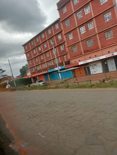 This media file captures a street scene in Karatina, Kenya, under an overcast sky, suggesting a cloudy day. The image is taken at a slight tilt, possibly from a moving vehicle.
The central subject is a prominent multi-story building, constructed with red bricks, which dominates the mid-ground. This building appears to be commercial or residential, featuring numerous rectangular windows across its multiple floors. The ground floor of this building houses several small businesses or shops, some with their roll-down shutters closed in various colors like blue and orange, while others appear open.
Visible text on a sign above one of these ground-level establishments clearly reads "BRASH AUTO ZONE." Another sign to its left shows partially legible text, possibly "SURP" or a similar word.
In the foreground, a paved road stretches across the lower half of the image. The road surface shows signs of wear and tear, with numerous cracks visible. To the left of the road, a white pickup truck is parked on the unpaved shoulder. Beyond the road and vehicle, some green foliage and trees are visible in the distance, indicating a semi-urban environment. Utility poles or streetlights are also present along the side of the road, leading up to the building. No people are directly visible, and no specific activity is taking place other than the general street view.