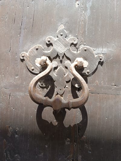The image shows a close-up of a weathered, brown wooden door with an ornate iron door knocker. The knocker features a decorative backplate with floral and geometric elements, and a curved handle that casts a shadow on the lower part of the door. The door itself is aged and shows signs of wear, with visible wood grain and cracks. The image was taken in Mezzana Sant'Antonio, Italy.