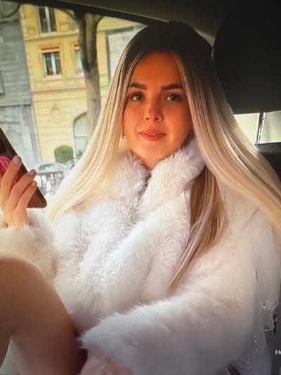 The image shows a medium shot of a young woman with long, blonde hair, wearing a white fur coat, sitting in a car. She is holding a pink phone in her left hand. The background appears to be an urban setting with buildings visible through the car window. The image is pixelated and of low resolution. There is no indication that this image is related to the city of Chorzów, Poland.