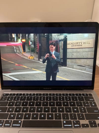 A MacBook Air laptop displays a scene from a show or movie. The screen shows a man in a suit standing on a street corner in front of a building with "Rendipity Hill Luxuryhouse" displayed. The keyboard of the MacBook is dusty and shows typical key arrangements. The laptop is on a light-colored surface.