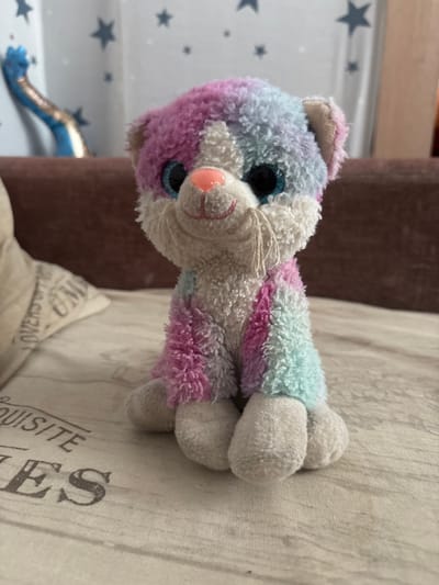A close-up, eye-level shot features a stuffed cat with pastel tie-dye fur in shades of pink, purple, blue, and white. The cat is sitting upright, facing the camera, with large, sparkly blue eyes and a small pink nose. Its white whiskers are visible. The cat is positioned on a textured, light-colored surface, possibly a blanket or tablecloth, with a subtle pattern. Behind the cat, out of focus, is a brown sofa and a pale fabric object with text that appears to say "QUISITE" and "ES." In the background, a sheer curtain with dark grey star patterns is visible. To the left, a blurry blue object with a golden element is partially visible. The scene is illuminated by ambient light, casting soft shadows. The location is unknown, but the query asks to include details about Клейнікаўскі сельскі Савет, Belarus, which are not present in the image.