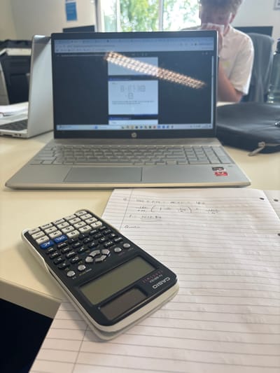 The image shows a desk in a room in Lancaster, United Kingdom, at coordinates 54.005, -2.79. A silver laptop is open with its screen displaying an internet browser window. A black calculator lies on a white desk. The calculator is positioned on top of lined paper with a handwritten math equation. There is a notebook behind the calculator. An out-of-focus individual in a white T-shirt sits on the left side of the image, seemingly in the background. The scene is likely part of a study area or a classroom. The laptop and calculator are the main subjects of the image.