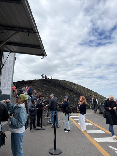 The image depicts a group of people standing on an asphalt surface with yellow and white stripes, likely a designated walkway or pedestrian crossing. The group is clustered near a gently sloping dark, volcanic-looking terrain, suggesting a location near a volcanic site. In the background, a cloudy sky dominates the upper portion of the frame. A metallic structure with an open panel and informative text is visible in the foreground. The location can be identified as near Mount Etna, given the volcanic terrain and the fact that Nicolosi is a common access point to the mountain.