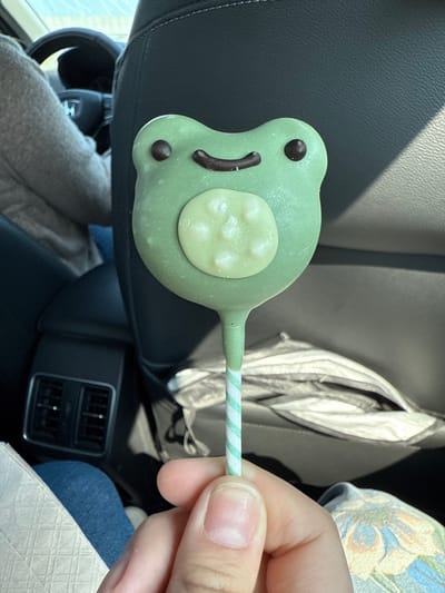 A hand holds a green frog-shaped lollipop with a green-and-white striped stick. The lollipop has brown eyes and a brown mouth. A light-colored circle with white spots is on the frog's body. The background includes a car seat, the back of the front seat, and a blue-jeaned leg.