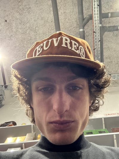 An individual is depicted in a close-up, low-angle indoor shot, positioned in Joinville-le-Pont, France. The male subject has fair skin, dark curly hair, and wears a brown baseball cap with "EUVRE®" embroidered in white capital letters on the front. He is also wearing a dark grey turtleneck sweater. The subject looks directly at the camera with a neutral expression. The background features an industrial-style ceiling with exposed pipes and a perforated metal cable tray. Below this, white shelving units are visible, displaying numerous packaged items, including various small, rectangular boxes in metallic and green finishes, suggesting products like fragrances or cosmetics. A handwritten label, partially obscured on a shelf to the right, appears to read "CALVIN K". A bright light source is present in the upper left portion of the image.