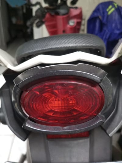 The image shows the rear of a motorcycle, focusing on the taillight. The taillight is red, oval-shaped, and surrounded by a dark gray or black plastic housing. The seat is visible above the taillight and appears to be made of a dark, textured material. The motorcycle body is a combination of white and gray. A red object is visible behind the seat.