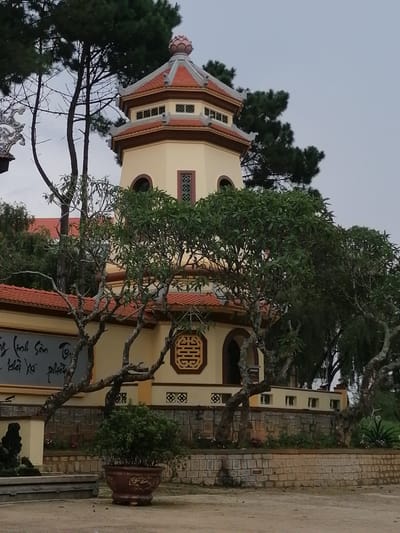 This image captures a view of a pagoda in Da Lat, Vietnam. The pagoda, a tall octagonal structure, is painted a pale yellow with a red tiled roof. It features several windows with intricate latticework and decorative elements on its facade. The pagoda is surrounded by lush green trees, some with gnarled branches, and a stone wall with patterned openings. In the foreground, a large potted plant sits on the ground, adding to the serene atmosphere. The overall scene suggests a peaceful and culturally rich environment, typical of the spiritual sites found in Da Lat.