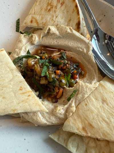 A close-up shot shows a plate of hummus topped with a dark relish, surrounded by pieces of pita bread. A silver spoon rests partially on the plate, near the edge of the hummus. The relish appears to contain chopped vegetables and herbs, glistening with oil. The pita bread pieces are cut into triangular shapes. The plate has a speckled pattern. The location appears to be the city of Oak Brook, United States of America.