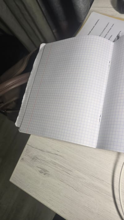 This media file is a close-up, high-angle shot of an open notebook with graph paper, resting on a light-colored, wood-grain textured surface. The notebook is positioned at an angle, with the right side of the page facing upwards. A portion of a white cable is visible at the top right of the image, suggesting it might be connected to a device or charging. The background is blurred, showing dark, indistinct shapes that could be furniture or other objects.
The notebook pages have faint blue grid lines and a red horizontal line near the bottom of the visible page. There are some staples holding the pages together, visible on the left side of the open notebook.
There is visible text on the left-hand page of the notebook, written in a dark font. The text appears to be in Russian and includes phrases like "еский анализ данных" (possibly referring to data analysis), "в условиях рыноч" (in market conditions), "системы пока-" (system shows), and "ного прироста" (growth). The text is oriented such that it reads upwards from the bottom of the page.
The lighting suggests an indoor setting, possibly an office or study area, with artificial light. The overall impression is one of a workspace with a focus on a notebook containing written information. No people are visible in the frame. The scene is static and appears to be a still photograph rather than a video. The texture of the table and the paper are prominent details. The context of Ukhta, Russia, is provided but not visually evident within the image itself.