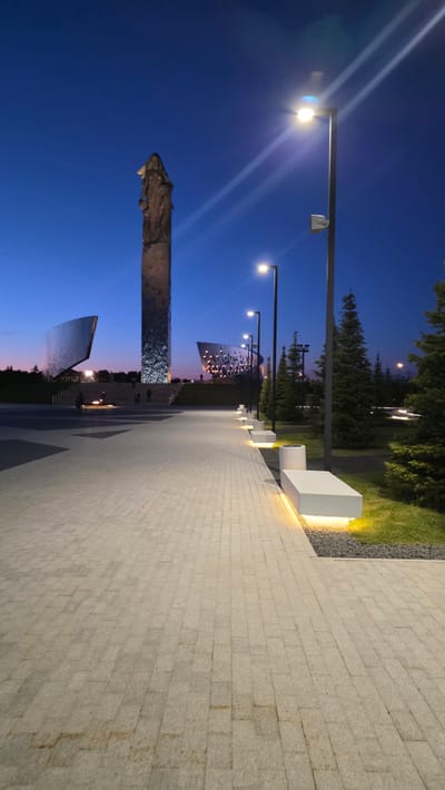 The image shows a paved walkway in Zaitsevo (Verevskoe S/P), Russia, at dusk or night. The walkway is constructed of light-colored rectangular stones, laid horizontally. Parallel to the walkway are regularly spaced benches or light fixtures. To the left of the walkway is a monumental sculpture or structure reflecting the evening sky, consisting of a central high structure with a statue or figure at its top. There are two curved mirror-like structures. Streetlights illuminate the walkway, casting bright light. Trees and vegetation border the walkway on the right, with a section of grass between the walkway and the greenery. The sky transitions from blue to a subtle pink or orange hue near the horizon. There are people walking along the walkway.