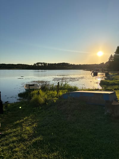 This image depicts a serene sunset scene at a lake, possibly located in or near Chikino, Russia. The sun, a bright orb, is setting on the right side of the frame, casting a warm golden glow across the water and sky. The sky transitions from a clear blue at the top to a lighter yellow near the horizon. In the foreground, the grassy bank slopes down towards the lake. A small, white boat with a green interior is partially submerged at the water's edge, grounded on the shore. Further along the bank, an overturned blue boat rests on the grass, with a tree stump to its right.
The lake itself is calm, with ripples on its surface reflecting the sky and the distant treeline. Scattered patches of aquatic vegetation are visible in the water. Across the lake, a dense forest lines the horizon, silhouetted against the bright sky. On the right side of the lake, a wooden pier extends into the water, with a few people standing on it. The pier has railings and steps leading down to the water. The overall atmosphere is peaceful and tranquil, characteristic of a summer evening.