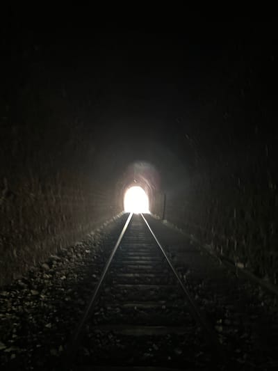 An interior view from inside a dark train tunnel shows a bright light at the end of the tunnel. Railroad tracks lead into the tunnel from the foreground, with ballast and gravel on either side of the tracks. The tunnel walls are rough and textured, appearing to be made of stone or brick. The scene is dimly lit except for the bright illumination emanating from the tunnel's exit. The perspective draws the viewer's eye towards the light, suggesting a journey or an escape from darkness.