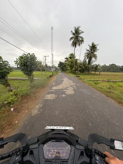 The image depicts a first-person perspective from a moving motorcycle on a wet, paved road in Keudekarieng, Indonesia, under an overcast sky. The black motorcycle's dashboard, handlebars, and a portion of the digital display are visible in the foreground, all covered in raindrops. The display shows "123 Km/h," a time of "6:28," and "245 12" (likely odometer/trip meter). A white license plate, "BG 4408 NAM," is mounted above the dashboard. A hand is visible gripping the right handlebar.

The asphalt road, exhibiting areas of wear and lighter patches, stretches straight into the distance, showing motion blur indicative of travel. To the left of the road, green fields, likely rice paddies, are visible beyond a strip of grass and small trees. Overhead power lines traverse the sky, supported by a wooden utility pole. A prominent, tall, lattice-structured cellular tower stands further down the road on the left, with a building partially visible behind it. To the right of the road, dense tropical vegetation, including several tall palm trees and other broadleaf trees, borders the path, with more green fields visible beyond. The sky is uniformly gray and overcast.