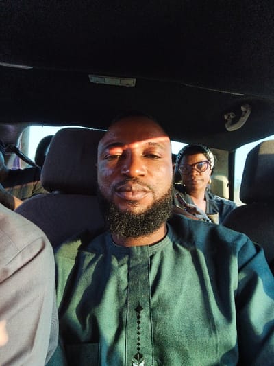 This close-up selfie captures two individuals inside a car, likely in Aboro, Nigeria. The foreground is dominated by a man with a dark beard and short hair, looking directly into the camera with a neutral expression. He is wearing a dark green traditional garment featuring intricate black embroidery down the center placket. A strong, warm ray of sunlight strikes his forehead and face, creating a distinct highlight and shadow effect, suggesting a low-angle sun, possibly late afternoon or early morning.
Behind the man, a woman is visible, wearing eyeglasses and a patterned headwrap. She also has a neutral expression and appears to be looking forward, not at the camera. She is wearing a dark top and holds a small, dark object in her hands. The car's dark interior, including headrests and ceiling, is visible around them. A partial view of another person's shoulder can be seen on the far left. The overall impression is one of people traveling together in a vehicle during daylight hours. No visible text is present in the image.
