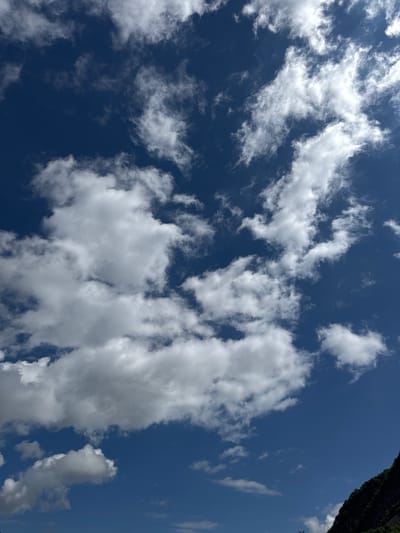 The image depicts a clear, deep blue sky filled with a variety of white clouds. Some clouds are thin and wispy, appearing as streaks across the sky, while others are more voluminous and puffy. In the bottom right corner, a dark green, tree-covered mountainside is visible, angled diagonally into the frame. The lighting suggests it is daytime, with the sun likely positioned above and to the right, illuminating the clouds and the mountain. There are no people or discernable objects in the sky. The scene appears to be an outdoor natural landscape, possibly taken from a vantage point looking up at the sky and towards a mountainside.