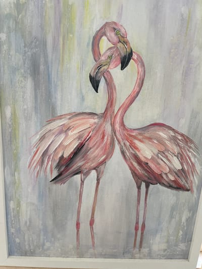 This is an oil painting of two pink flamingos standing close together, with their necks intertwined in a heart shape. The background is abstract with vertical brushstrokes of grey, white, and muted colors like yellow and purple. The flamingos are painted in varying shades of pink, with accents of white and some darker feathers. Their eyes are visible and appear yellow. The painting is framed in white. There is a signature in the bottom right corner, which is difficult to read. The painting is displayed indoors, likely in a home or gallery setting. There is no information available about its location in Moscow, Russia.
