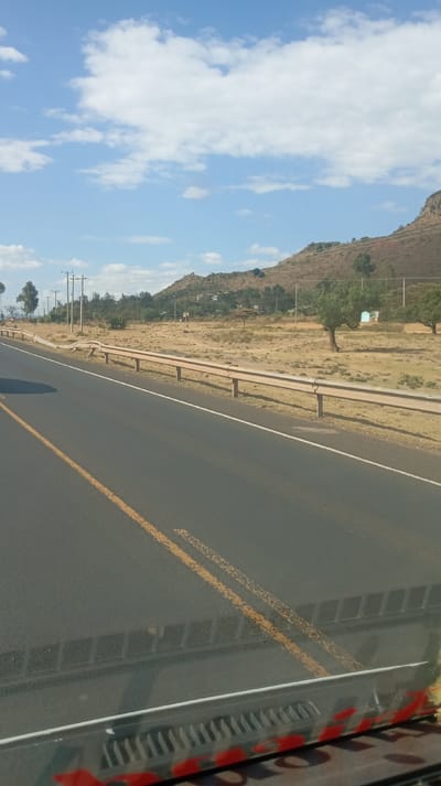 A sun-drenched highway stretches into the distance under a partly cloudy sky. To the right, a hillside with sparse vegetation and scattered structures rises above the roadside. A rusty metal guardrail runs parallel to the road, separating it from the grassy terrain. Telephone poles line the area. The photo appears to be taken from inside a vehicle, with part of the dashboard and windshield visible at the bottom.