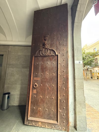 The image shows a large, ornate wooden door leaning against a stone archway in Mataró, Spain. The door is made of dark wood and is studded with small metal rivets. It features a carved crest above a keyhole and a circular metal handle. To the left of the door, a metallic trash can sits on a tiled floor. The background reveals a street scene with buildings and trees, illuminated by daylight. A sign with the number "112" is visible on the stone pillar to the right of the door.