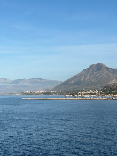 This vertical image depicts a panoramic coastal scene under a clear blue sky, identified as being located in or near the city of Sperone, Italy.
The lower half of the image is dominated by an expanse of deep blue, slightly rippled water, indicating a calm sea surface. Extending from the right mid-ground into the water is a long, curved stone breakwater, appearing light grey. A small, light-colored boat with a single occupant is visible on the water to the left of the breakwater's end. Another small, white boat is visible closer to the marina on the right.
Above the breakwater, the coastline reveals a densely developed urban area, forming a town or city. Numerous buildings, predominantly light-colored (white, cream, pale grey) with some red-tiled roofs, extend along the shore and up the lower slopes of the adjacent mountains. A prominent marina is visible behind the breakwater, crowded with numerous moored sailboats and yachts, identifiable by their white masts and structures. An elevated white structure, possibly a bridge or walkway, spans part of the marina. Towards the left of the town, a tall, slender, white industrial-style stack or chimney rises above the buildings.
The background is characterized by a range of mountains. A large, prominent, rugged mountain with dark grey and greenish-brown hues dominates the right side of the frame, showcasing visible rock formations and slopes. To the left, a series of more distant, lighter grey-blue mountains extends into the haze, with low-lying clouds or mist obscuring their bases. The sky above is a gradient of clear blue, transitioning from a slightly deeper blue at the top to a lighter hue near the horizon, with minimal atmospheric haze. No other discernible people, specific actions, or significant interactions are evident.
