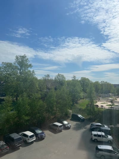 The image shows an elevated view of a street and surroundings in Volno-Nadejdinskoe, Russia. The sky is visible with blue patches and scattered clouds. A line of green trees separates the sky from the street. A row of parked cars lines the right side of the image. In the background, buildings and a playground are visible.