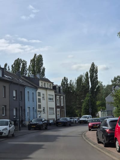 This image captures a street scene in Sanem, Luxembourg, on what appears to be a partially cloudy day. The perspective is from the asphalt road, looking forward as it curves gently to the right.
On the left side of the road, a row of multi-story residential buildings with varying facade colors is visible. From left to right, these include: a dark grey building, a light blue building, a cream-colored building with some glass block windows, and a dark brown building, all featuring multiple windows, some with external blinds. The roofs are dark and steeply pitched, equipped with dormer windows. A dark lamppost stands on the sidewalk in front of the blue building.
Several cars are parked along both sides of the street. On the left, partially visible in the foreground, is the rear of a white Renault Clio hatchback. Further ahead on the left, a black BMW X4 SUV is parked, displaying the Luxembourgish license plate "SB 8979". Further down the street on the left, a dark blue Audi SUV with license plate "JH 8483" is parked facing the viewer.
On the right side of the street, also parked, a red SEAT SUV with the Luxembourgish license plate "AG 1608" faces towards the viewer. Behind it, a white commercial van is parked, facing away. In the immediate foreground on the right, the rear section of a red hatchback car is partially visible. Further back, a black Mercedes-Benz CLA is also parked on the right.
The background on both sides of the street is dominated by numerous tall, slender deciduous trees, some appearing to be Lombardy poplars, suggesting a wooded area or tree-lined street. The leaves of these trees appear to be swaying or rustling, indicating a breeze. On the far right, a house with a dark slate roof is partially visible amidst the trees.
The sky above is light blue with scattered white cumulus clouds. Shadows are cast on the road surface, indicating the sun's position and suggesting mid-day lighting conditions. In the far distance on the right, behind the parked white van, two indistinct figures are visible walking on a path or verge.