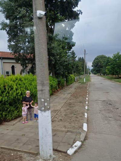 An overhead shot captures a dusty road stretching into the distance in the city of Dropla, Bulgaria. On the left side of the road, a dense, green hedge runs along the sidewalk. A woman in a black shirt stands with two children, a girl in a pink shirt and shorts and a boy in a beige shirt and blue shorts. The woman holds the girl's hand, and the girl holds the boy's hand. Both children are facing the camera. A utility pole stands to the right of the woman and children, and a security camera is attached to the pole. The road is paved and has white markings along its edge. The sky is overcast, and the surrounding environment is lush with trees.