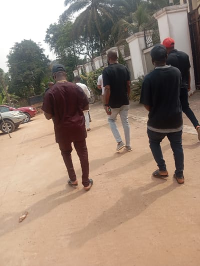 The scene depicts a group of approximately five to six adult males walking on an unpaved, reddish-brown dirt path in Okpatu-Ukehe, Nigeria. The men are largely viewed from behind, moving generally towards the right side of the frame. In the immediate foreground, a male wears a dark maroon traditional top and trousers with dark sandals, a black cap, and a visible gold-colored chain. To his right and slightly behind are other men: one in a white shirt and light trousers, another in a black t-shirt and light blue jeans with sneakers, another in a black t-shirt, dark trousers, and a black cap with dark sandals, and a man furthest right wearing a red cap, black shirt, and dark trousers. A single detached light-colored flip-flop lies on the ground in the lower left.

The environment features lush green vegetation, including tall trees and a prominent palm tree, behind a white building with a decorative black wrought-iron fence and gate structure. On the far left, two parked vehicles are partially visible: a silver SUV and a red sedan. Long shadows are cast on the path from the subjects, indicating direct sunlight. No direct interactions between the individuals are discernible.